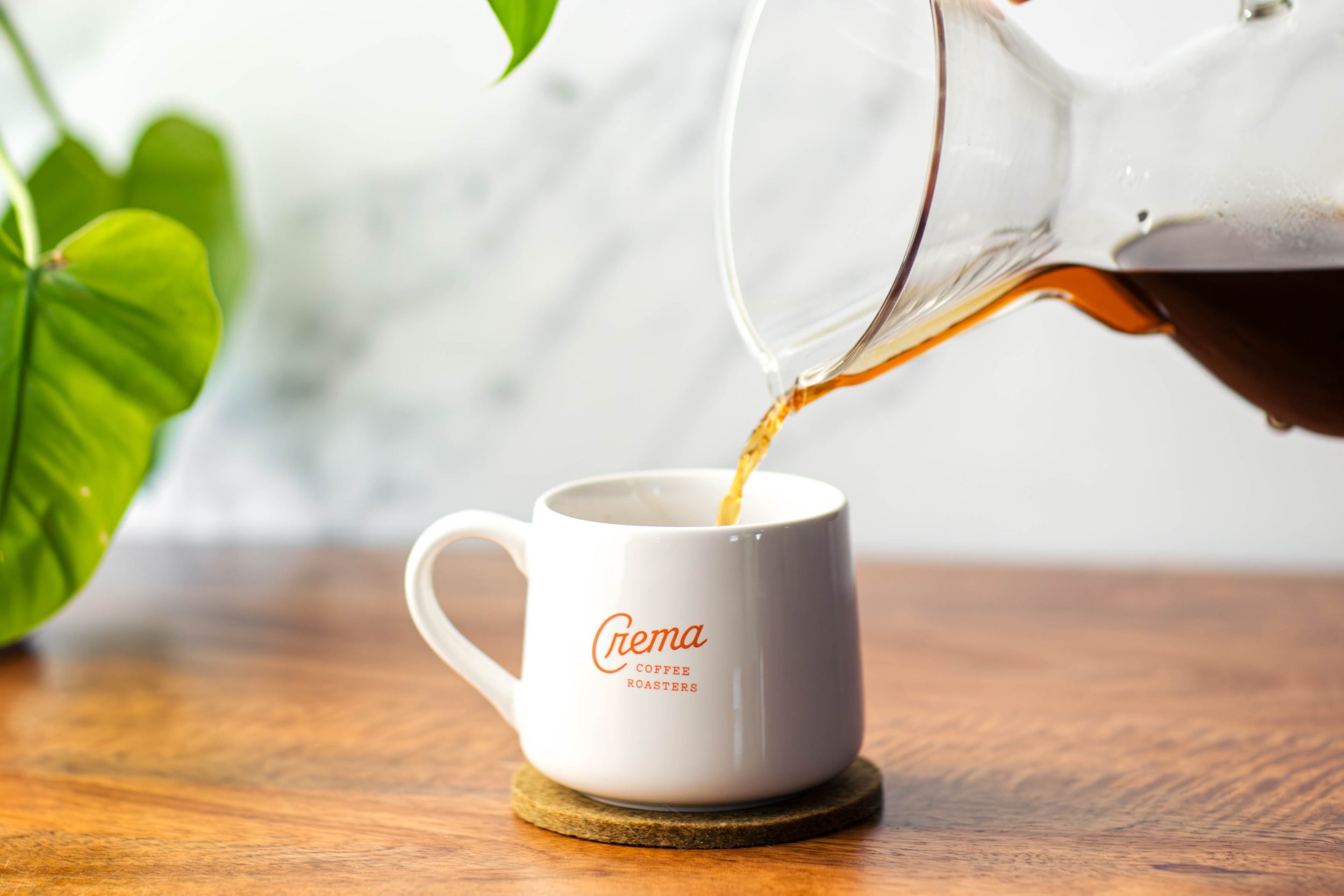 coffee being poured from a chemex into a white crema logo mug on a wooden tabletop