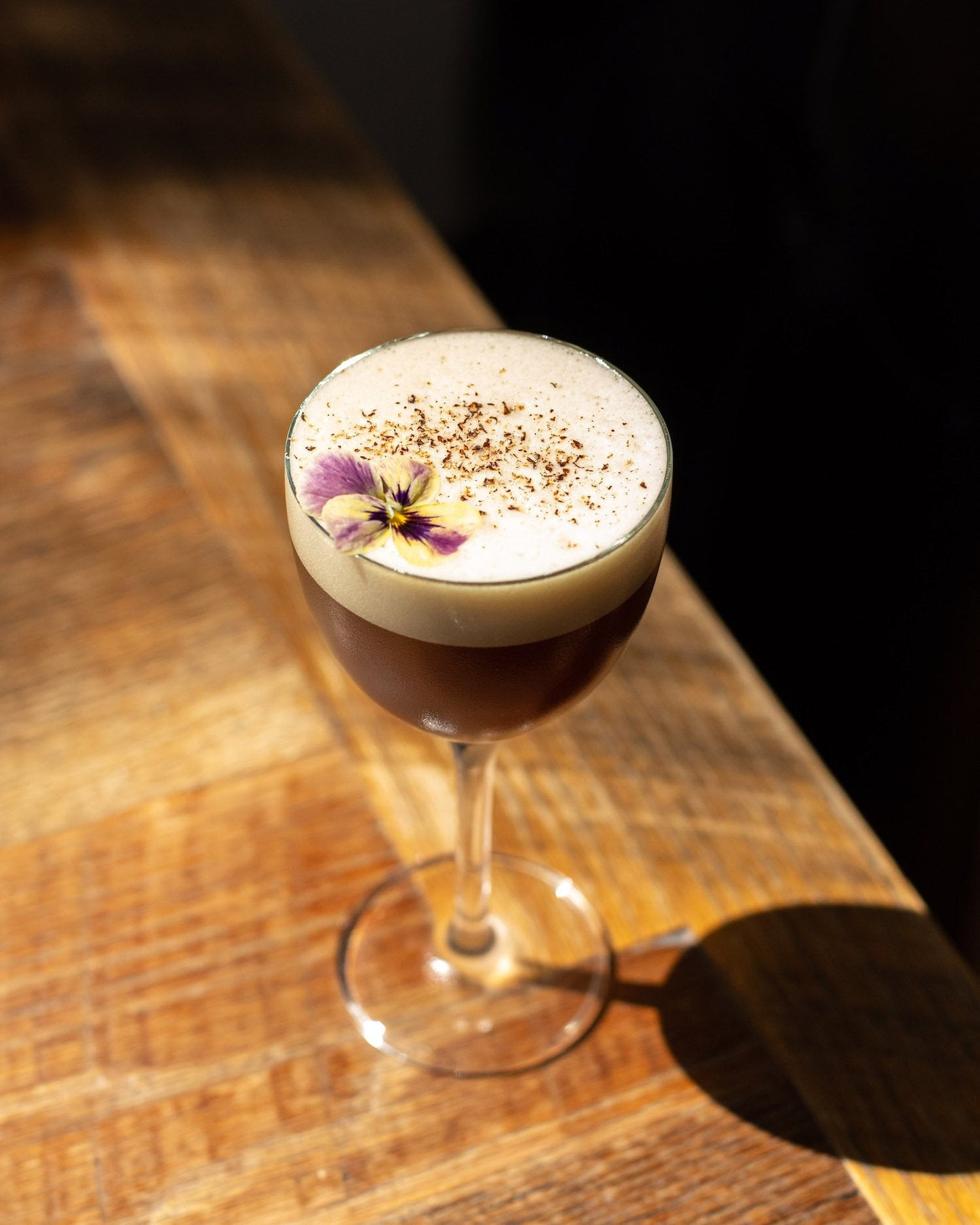 strange brew cocktail in coupe-like glass with a purple flower floating on top placed on a wooden table